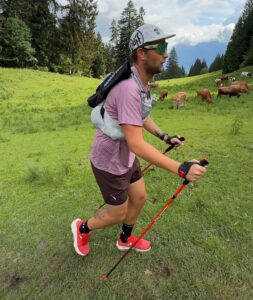 A trail runner moves across a green alpine meadow with poles.
