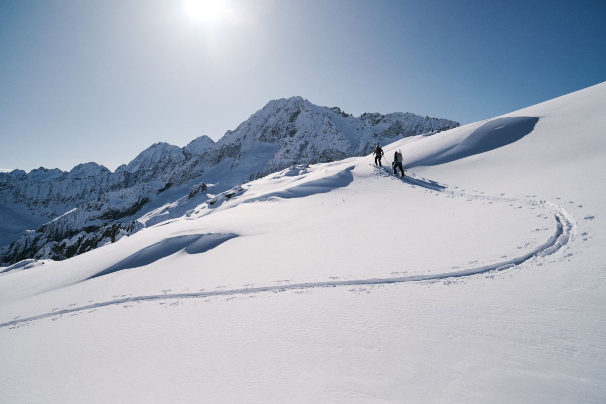 Two ski tourers ascending in sunshine with a mountain panorama in the background.