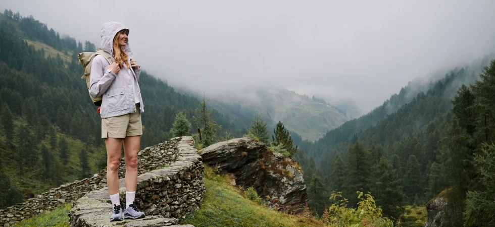 Frau mit Rucksack wandert bei Regen durch eine neblige Berglandschaft.