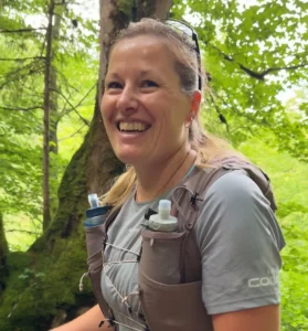 A smiling runner wears a hydration vest with soft flasks while moving along a forest trail.