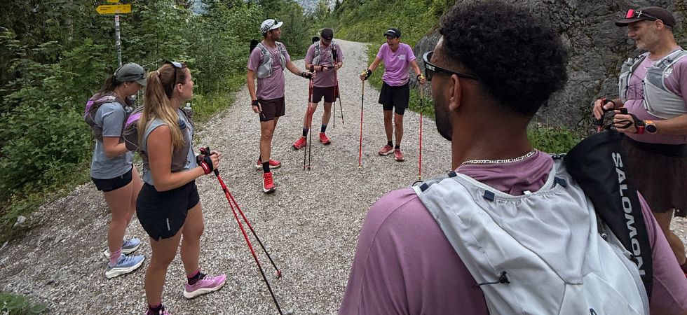 A group of trail runners wearing hydration vests and carrying poles discusses the route on a mountain trail.