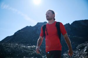 Hiker in a red shirt and backpack walking through a sunny alpine rock landscape.