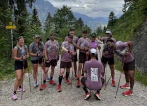 A group of trail runners poses with poles on a mountain path overlooking an alpine landscape.