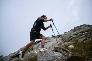 Läufer mit Trailrunning Ausrüstung steigt mit Stöcken über felsiges Gelände auf.