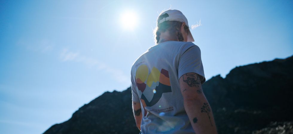 Person wearing a T-shirt and cap in bright sunshine against an alpine mountain backdrop.