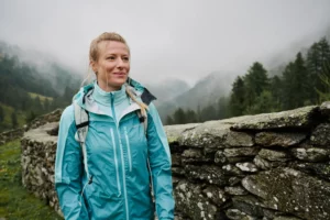 Woman in a turquoise rain jacket stands by a stone wall in a misty mountain landscape.