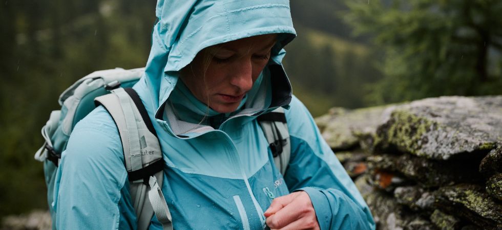 Woman in a turquoise rain jacket zips up her jacket in rainy mountain weather.