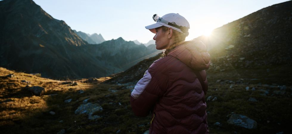 DE: Wanderin mit Stirnlampe steht bei Sonnenaufgang in den Bergen und blickt in die alpine Landschaft.