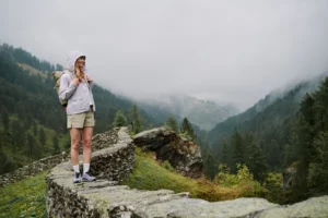 Hiker in a light rain jacket stands on a stone wall with a misty valley in the background.